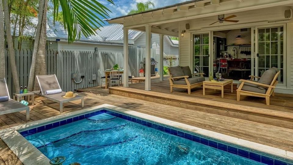 Private pool and patio area at a Key West vacation rental. The pool is surrounded by a wooden deck with lounge chairs and a table with chairs. Tropical plants and palm trees provide shade and privacy.